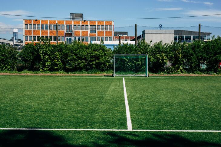 Tom fotballbane i et urbant område, med et fabrikkbygg i bakgrunnen.