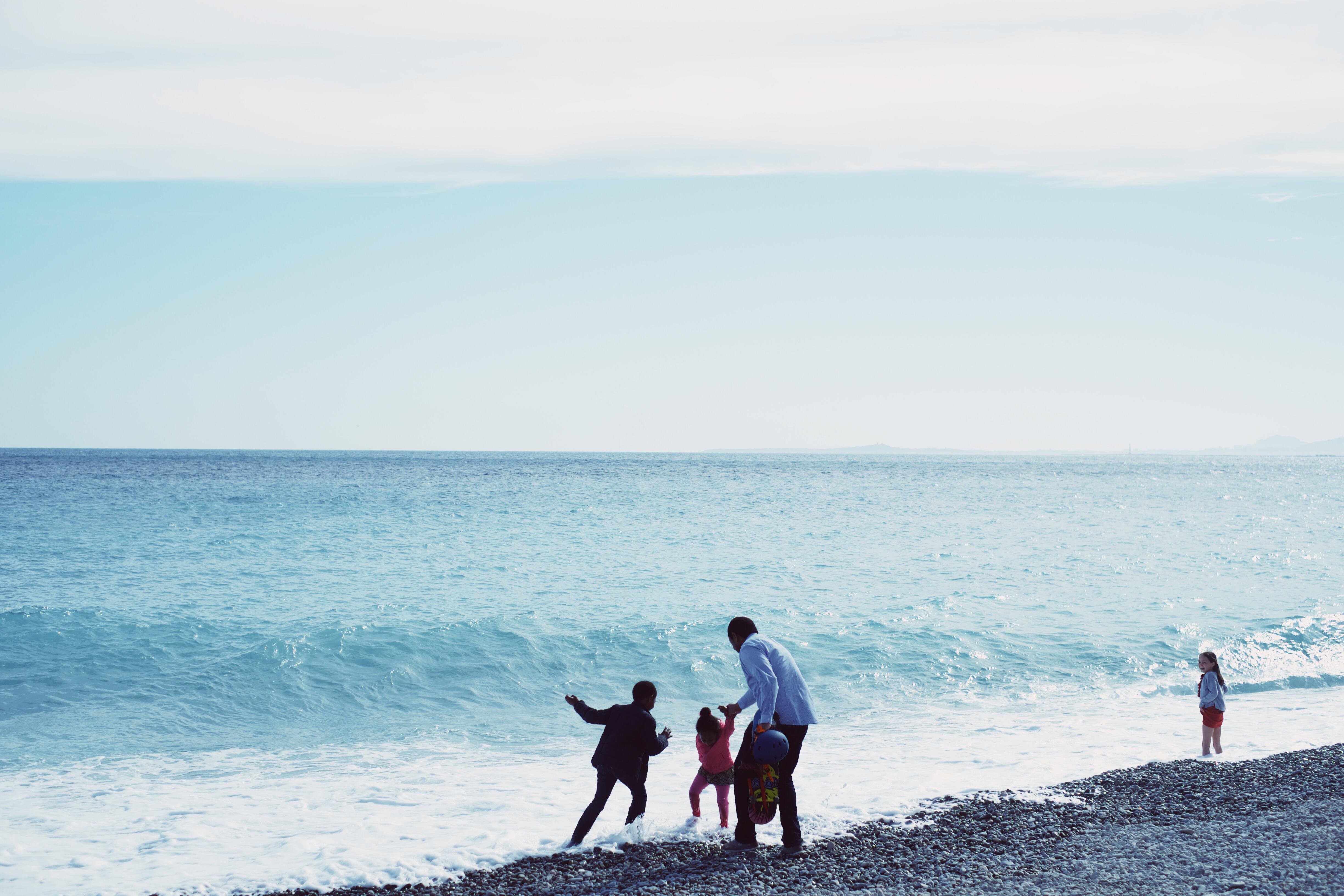 En voksen og to barn på en strand i Frankrike. Bølgene slår inn mot stranda. Et annet barn betrakter dem.